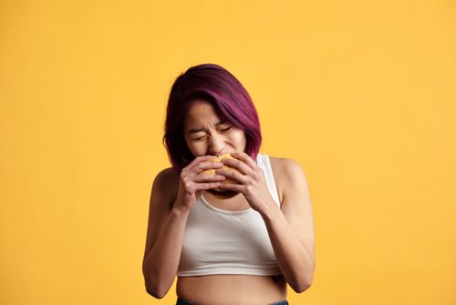 Free A vibrant studio shot of a young woman with purple hair savoring a burger against a yellow background. Stock Photo