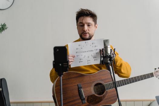 Man with acoustic guitar explains music theory online using smartphone and microphone.