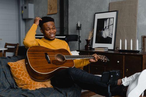 Free Young man in yellow sweater playing guitar on couch during video call session. Stock Photo
