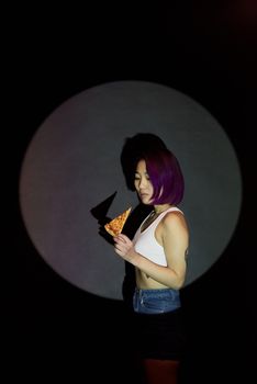 Asian woman with purple hair holding a pizza slice in spotlight.