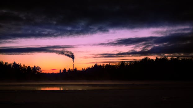 A serene sunset sky reflecting on water with industrial smokestacks silhouetted against the horizon.