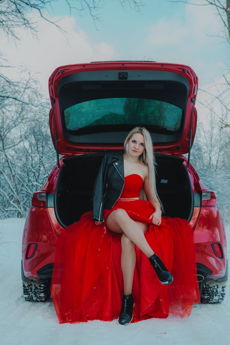 Woman In Red Dress Sitting In Trunk Of Car In Winter Forest