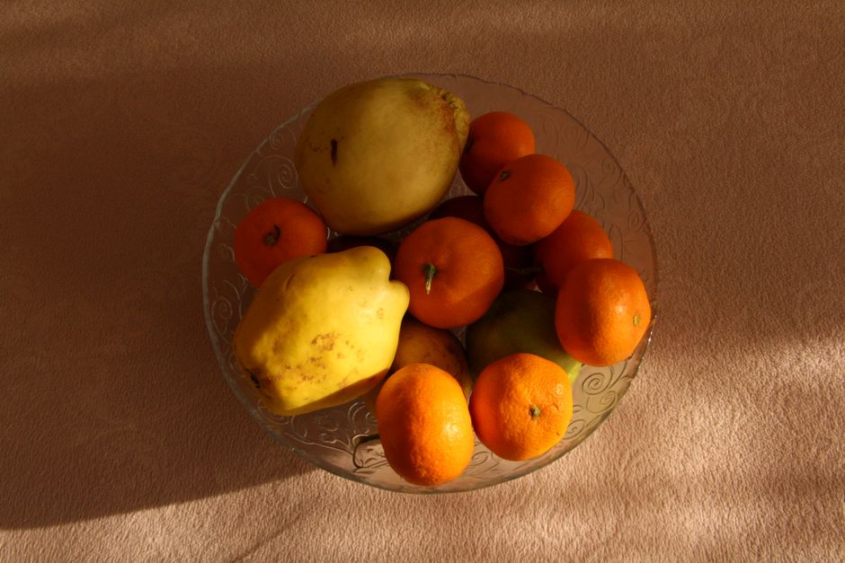 A variety of fresh fruits including quinces and oranges in a glass bowl under warm lighting.