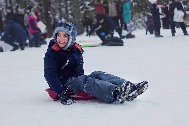 A Young Kid In Winter Clothes Smiling While Sitting On A Sled