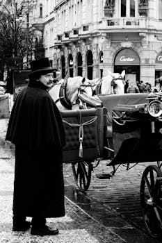 Black and white photo of a man in vintage attire beside horse-drawn carriages in a city street.
