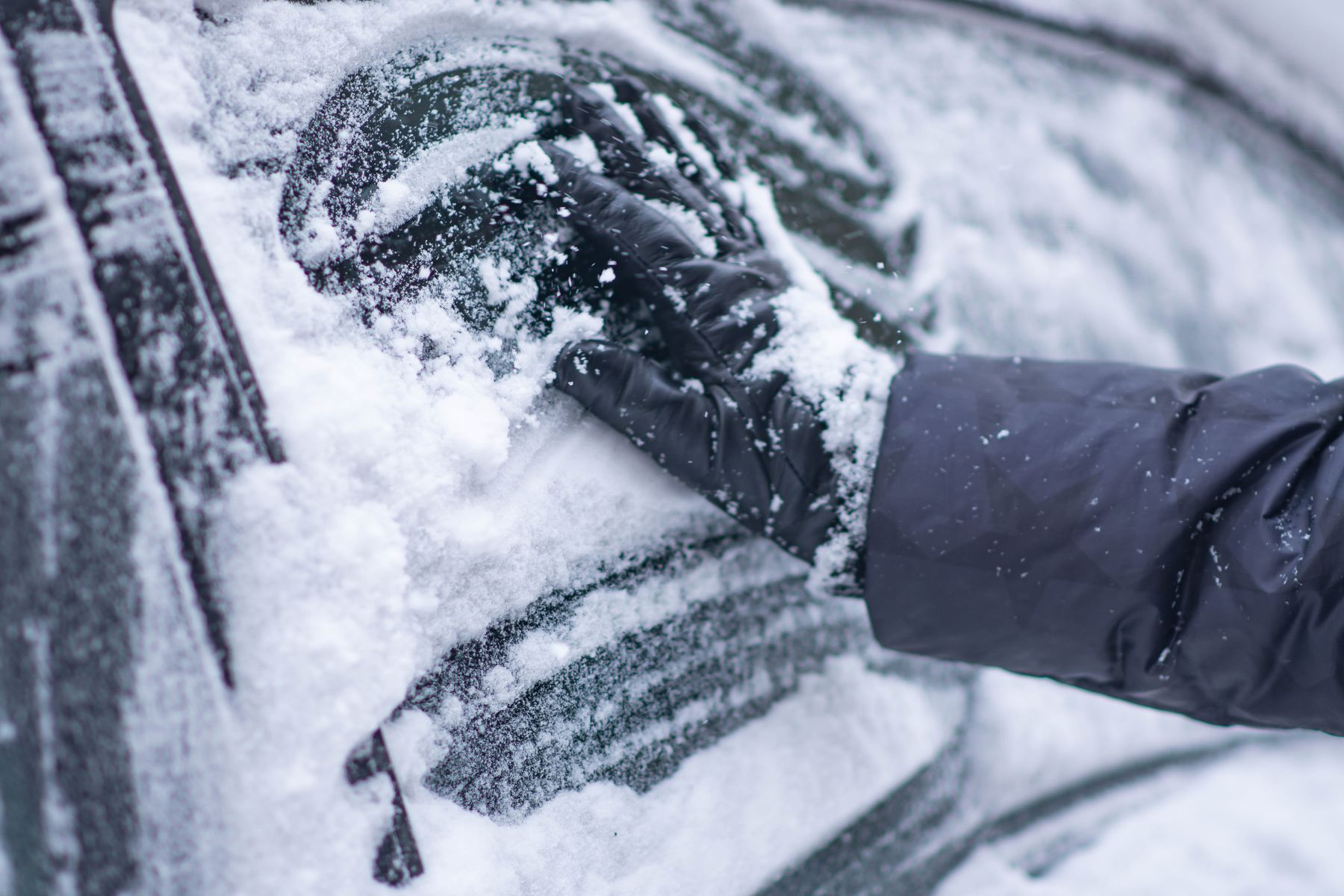 A gloved hand clearing snow from a car windshield on a winter morning