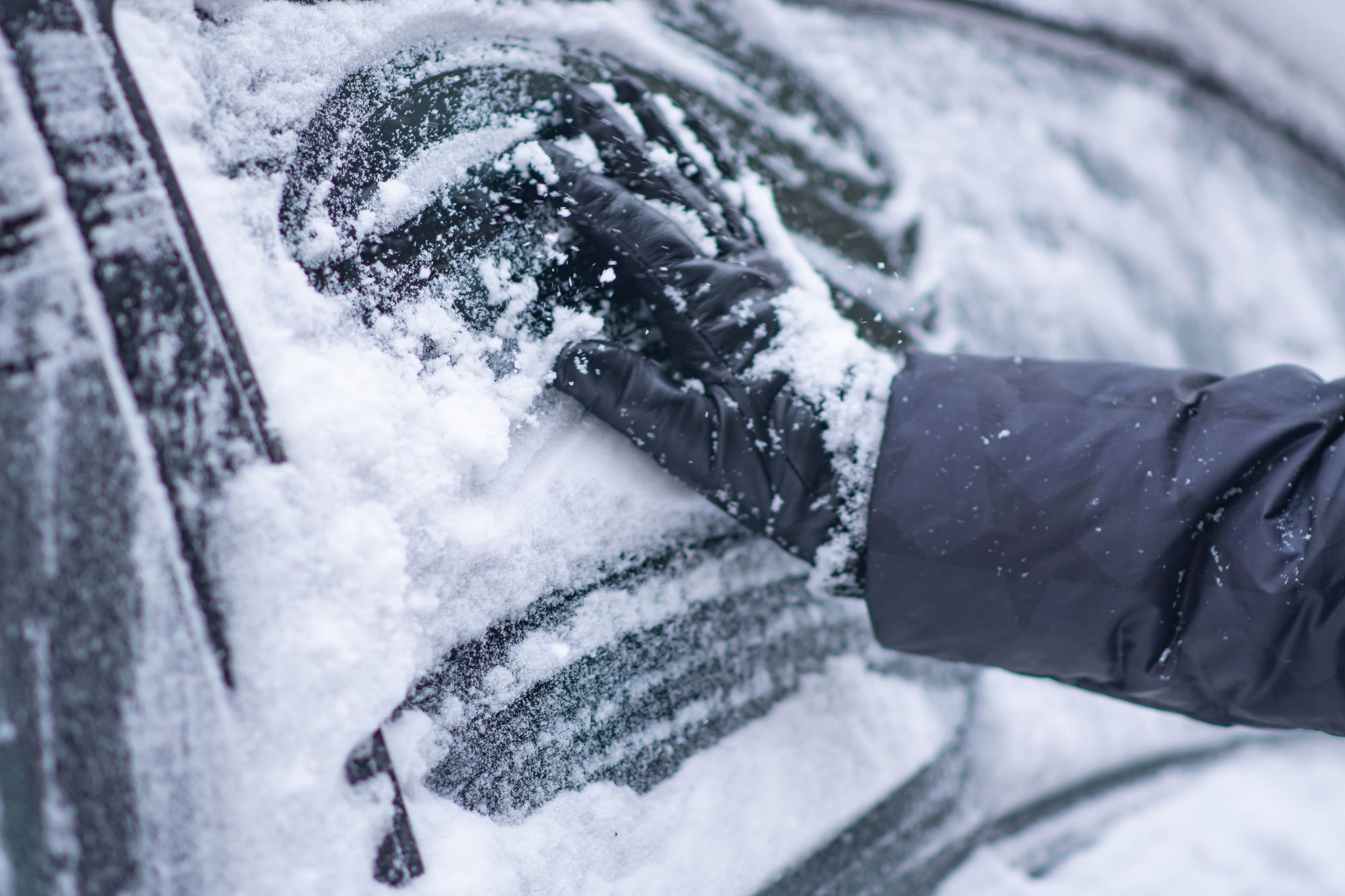 A gloved hand clearing snow from a car windshield on a winter morning