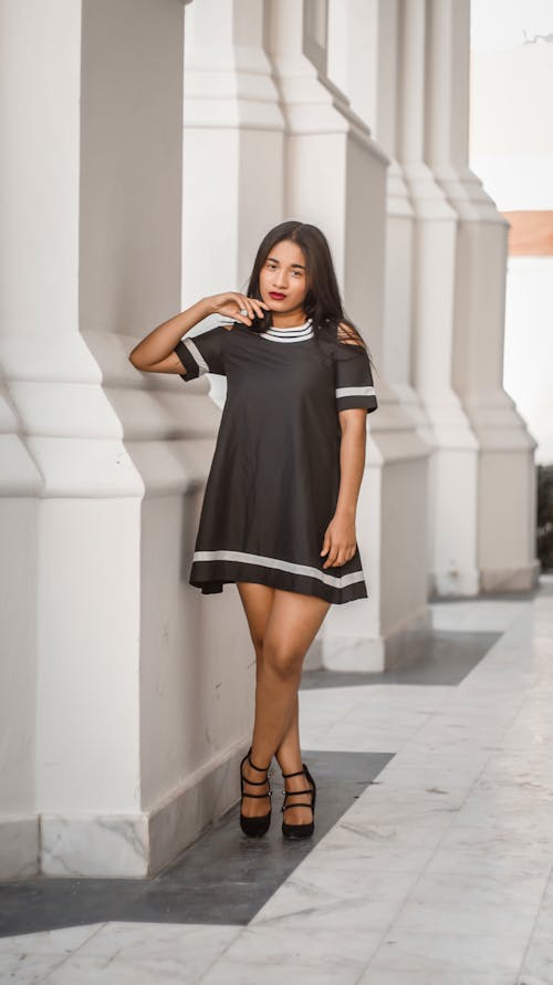 Woman in Black Dress Posing with Hand on Her Chin Beside White Concrete Wall