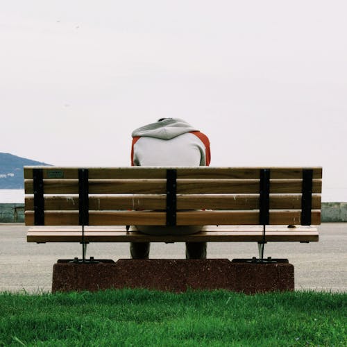 無料 昼間に茶色の木の公園のベンチに座っている灰色とオレンジ色のパーカーを着ている人 写真素材