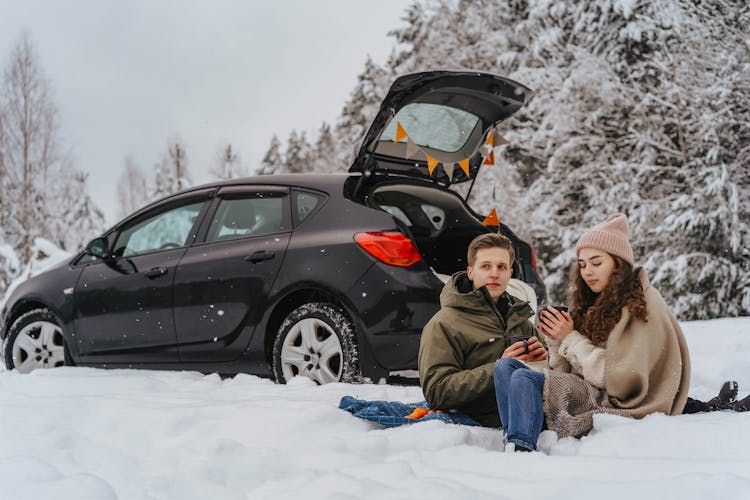 A Couple Sitting On A Snow Covered Ground Near The Black Car Parked Behind Them