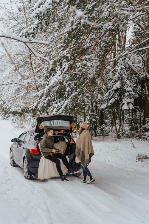 Free Couple sits by snow-covered car trunk in a romantic winter forest setting. Stock Photo