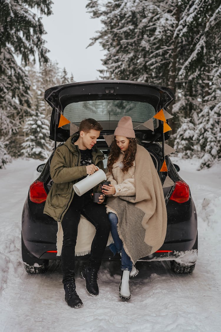 Man And Woman Sitting In A Car Trunk In Winter And Drinking Tea From A Thermos