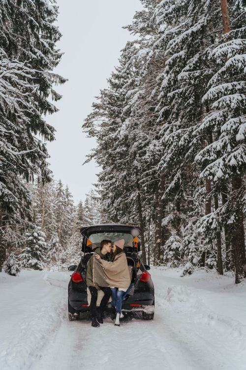 Free Couple sits in car trunk, wrapped in a blanket on a snowy forest road, embracing the winter season. Stock Photo