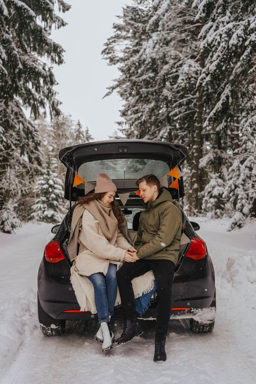 Free A couple sitting on a car trunk surrounded by a snowy forest, enjoying winter together. Stock Photo