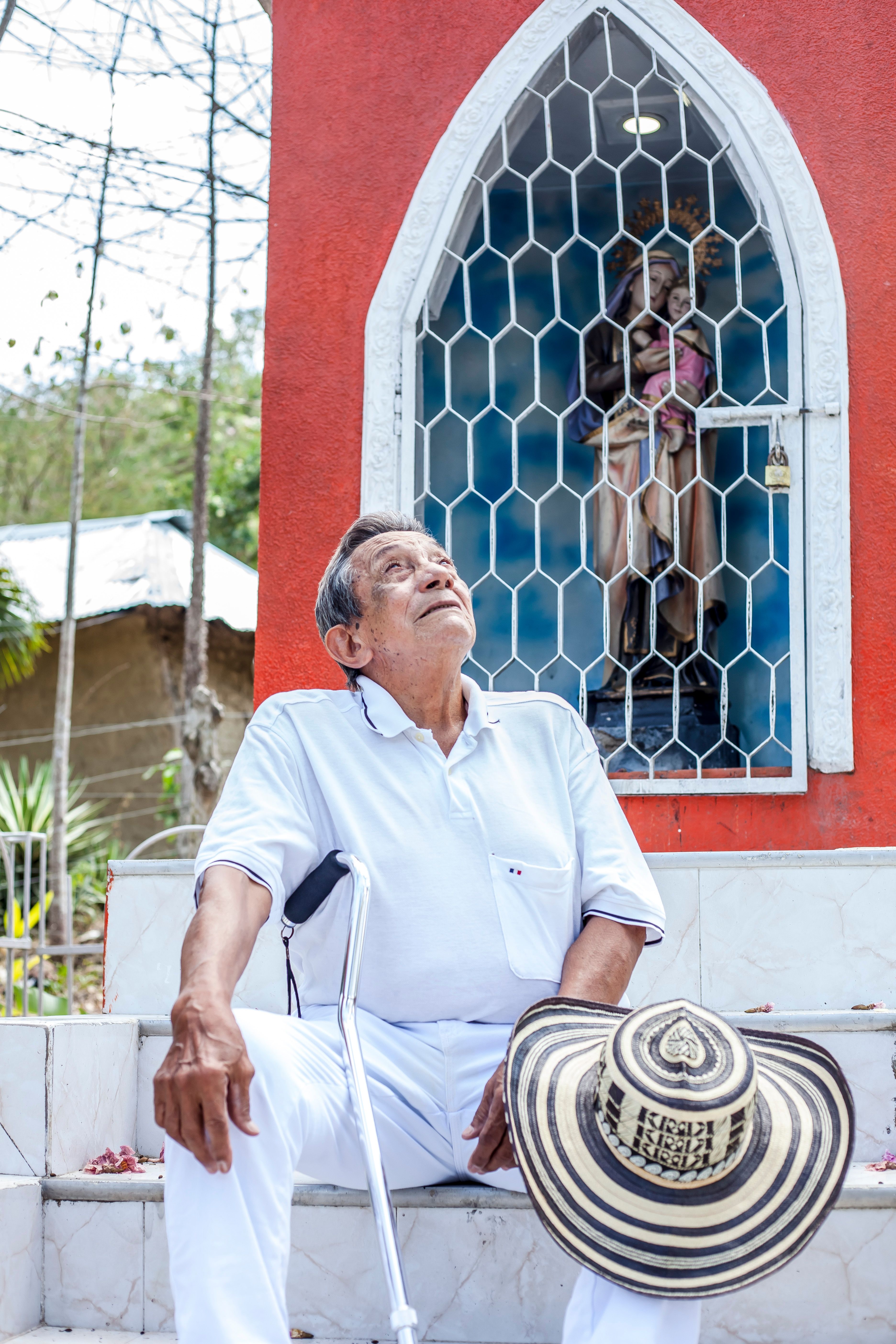 Senior man with cane and hat at a shrine in Ovejas, Colombia, contemplating.