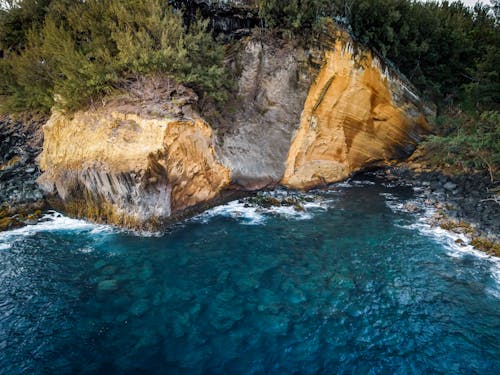 Rough rock with uneven surface and green trees placed on seashore with dense forest near calm turquoise water in nature