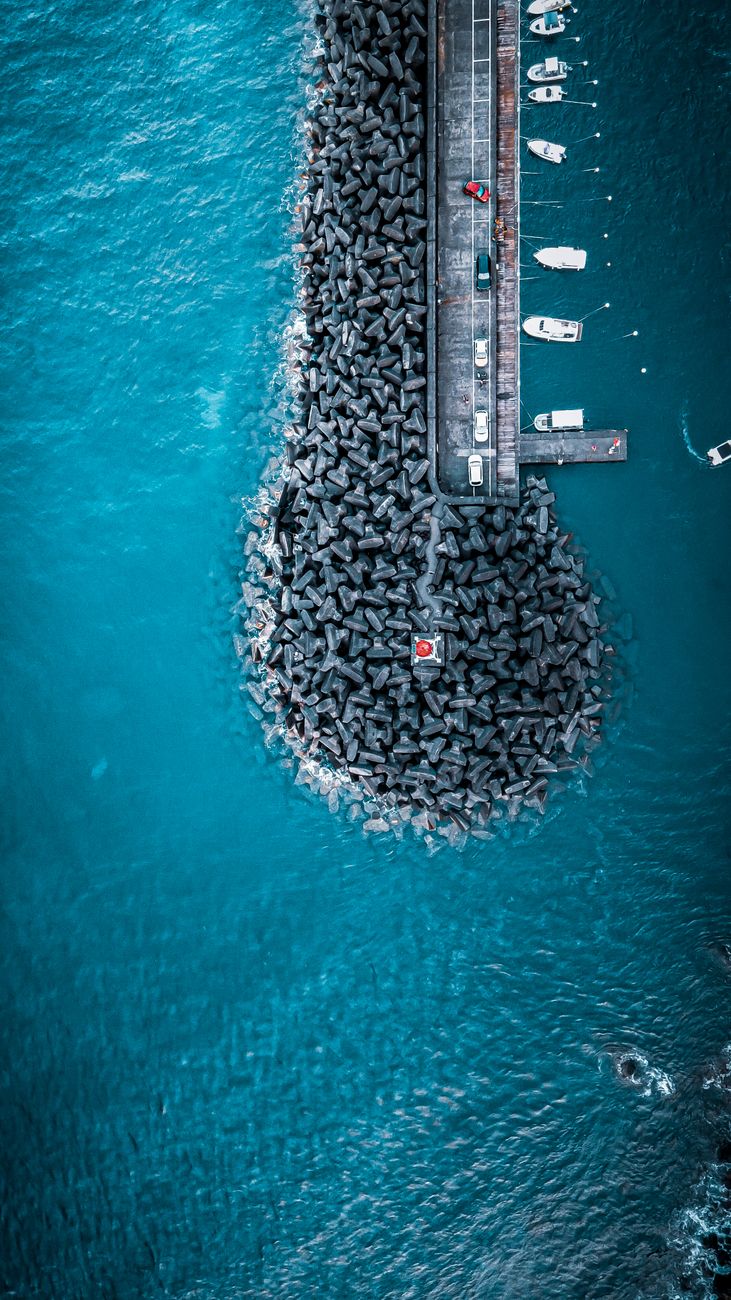 Drone view of waterfront with lighthouse with parked cars and modern yachts located among turquoise sea in nature in port