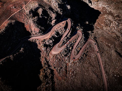 Drone view of cars riding on curvy roadway near tall rocky cliff in dry terrain with boulders in wild nature