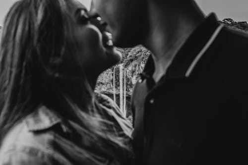 Black and white side view of anonymous boyfriend with girlfriend cuddling while standing in nature against rapid waterfall falling from rocky cliff