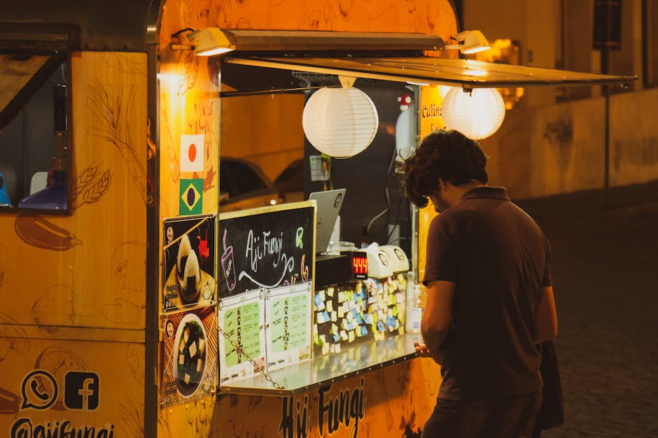 A food cart illuminated at night with a person waiting in front on a city street.