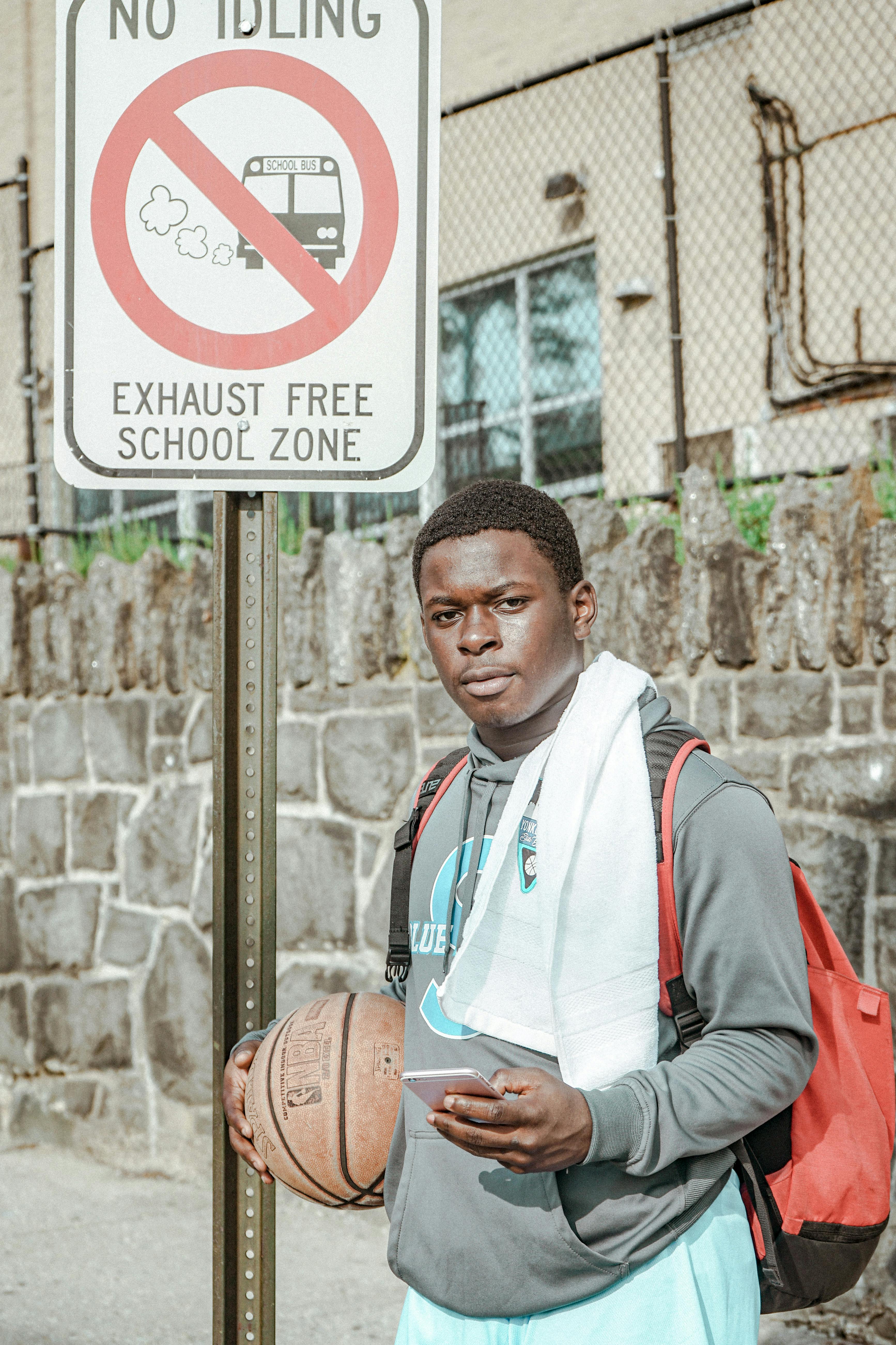 Young man holding basketball near a no idling sign in school zone.
