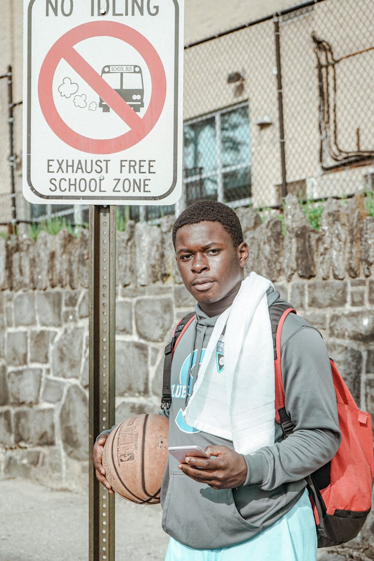 A Man Holding A Basketball And A Mobile Phone Standing Near The Street Sign