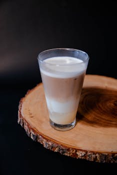 A tall glass of creamy latte sits on a rustic wooden coaster against a dark background.