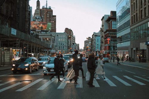 Group of people walking on pedestrian crossing on asphalt road with cars in city with residential buildings in evening time