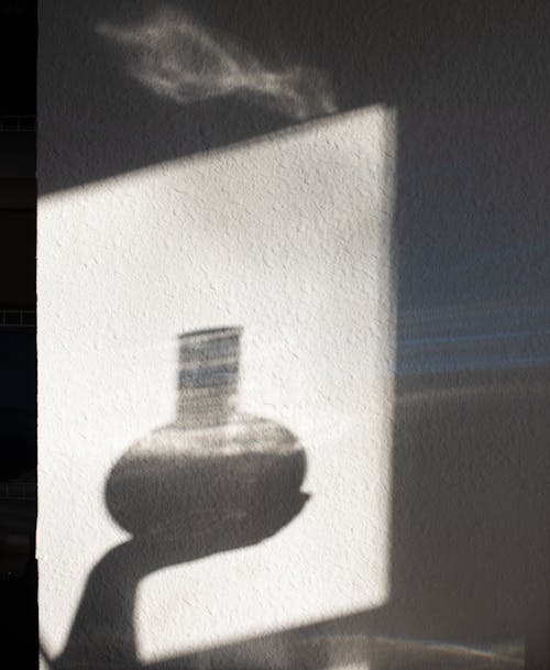 Shade of hand of crop unrecognizable person demonstrating round shaped bottle with shiny substance against white wall