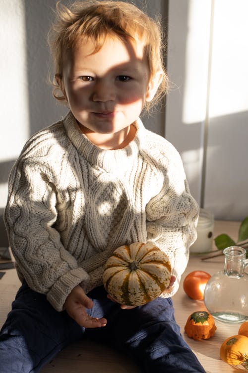 Adorable little child with blond hair in stylish knitted sweater sitting on wooden table with fresh small pumpkin in hand and looking at camera