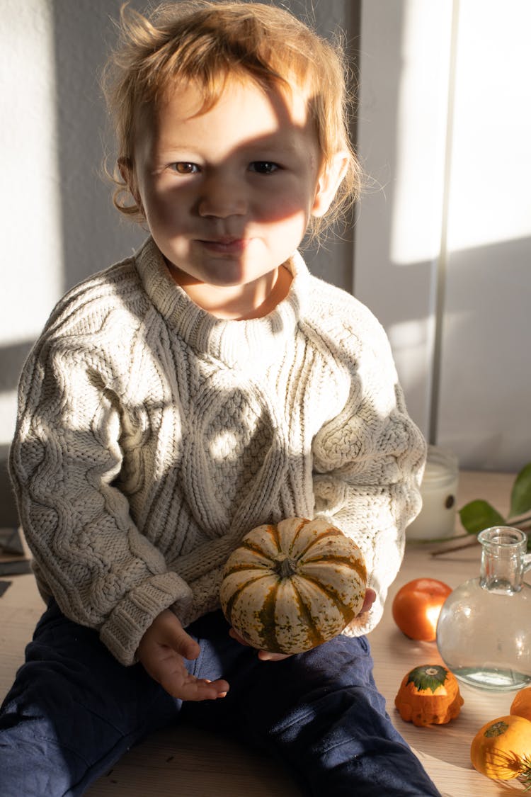 Cute Toddler With Pumpkin In Hand Sitting On Table In Sunlight