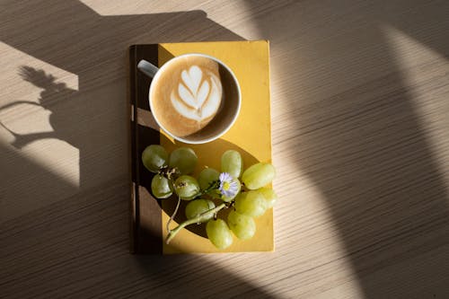 Free From above of fresh ripe green grapes and cup of fresh foamy cappuccino with latte art placed on book on wooden table in sunlight at home Stock Photo