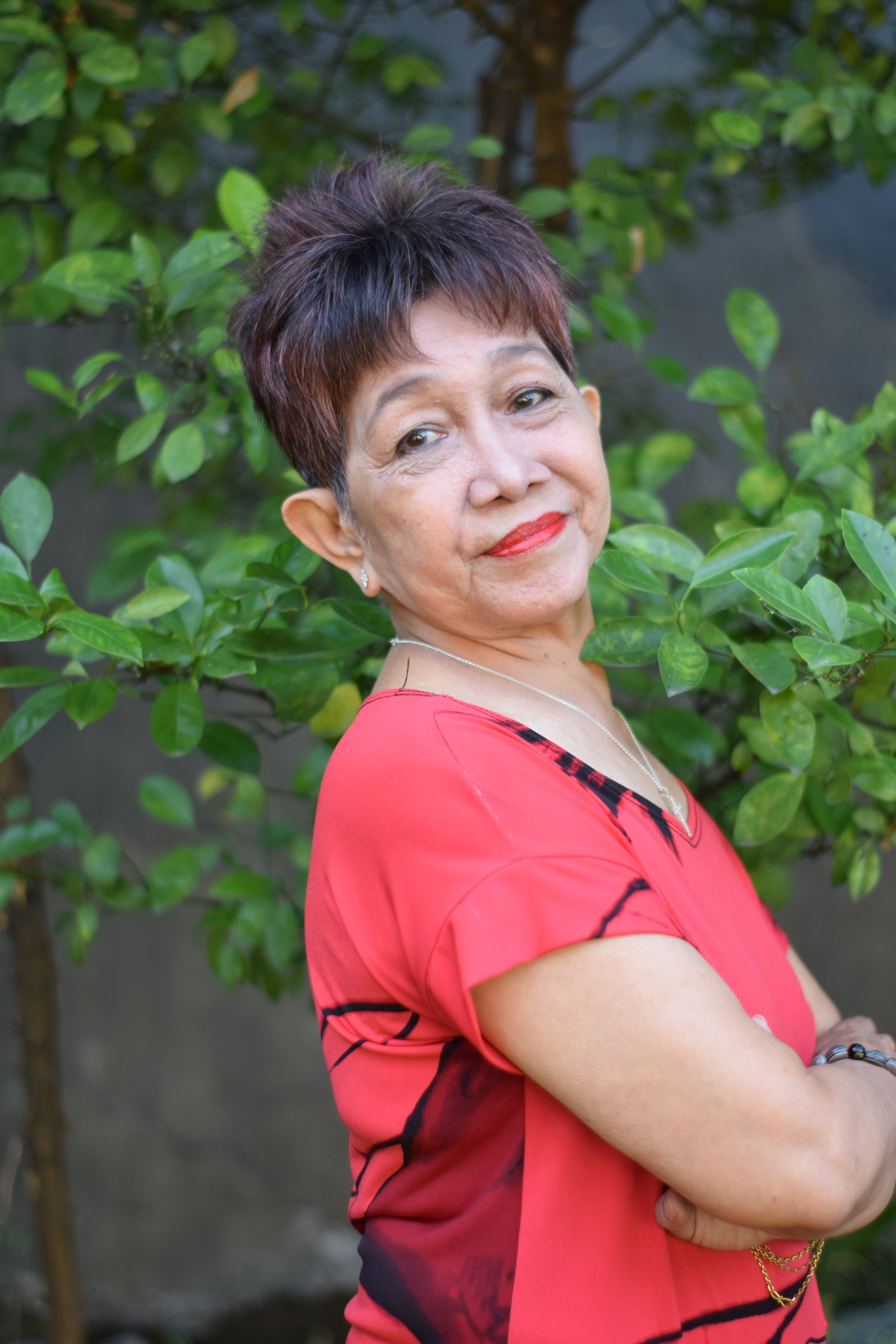 Smiling elderly woman in a red top stands relaxed outdoors by green foliage.