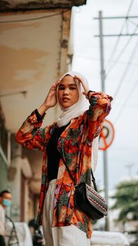 A young Muslim woman in a colorful outfit with a hijab poses confidently outdoors in a vibrant Cirebon street setting.