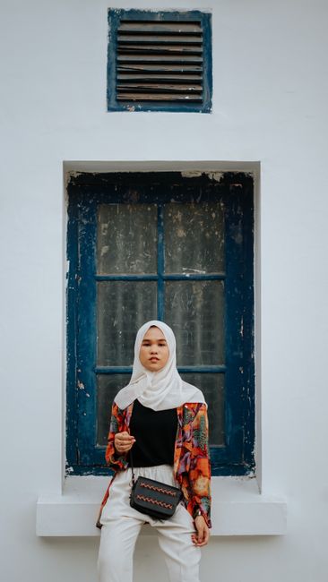 Portrait of a young woman in a colorful outfit and hijab, posing against a blue window backdrop.