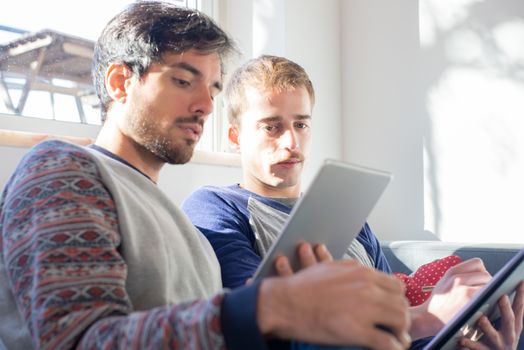 Two men sitting on a couch using digital tablets indoors with natural light, creating a cozy atmosphere.