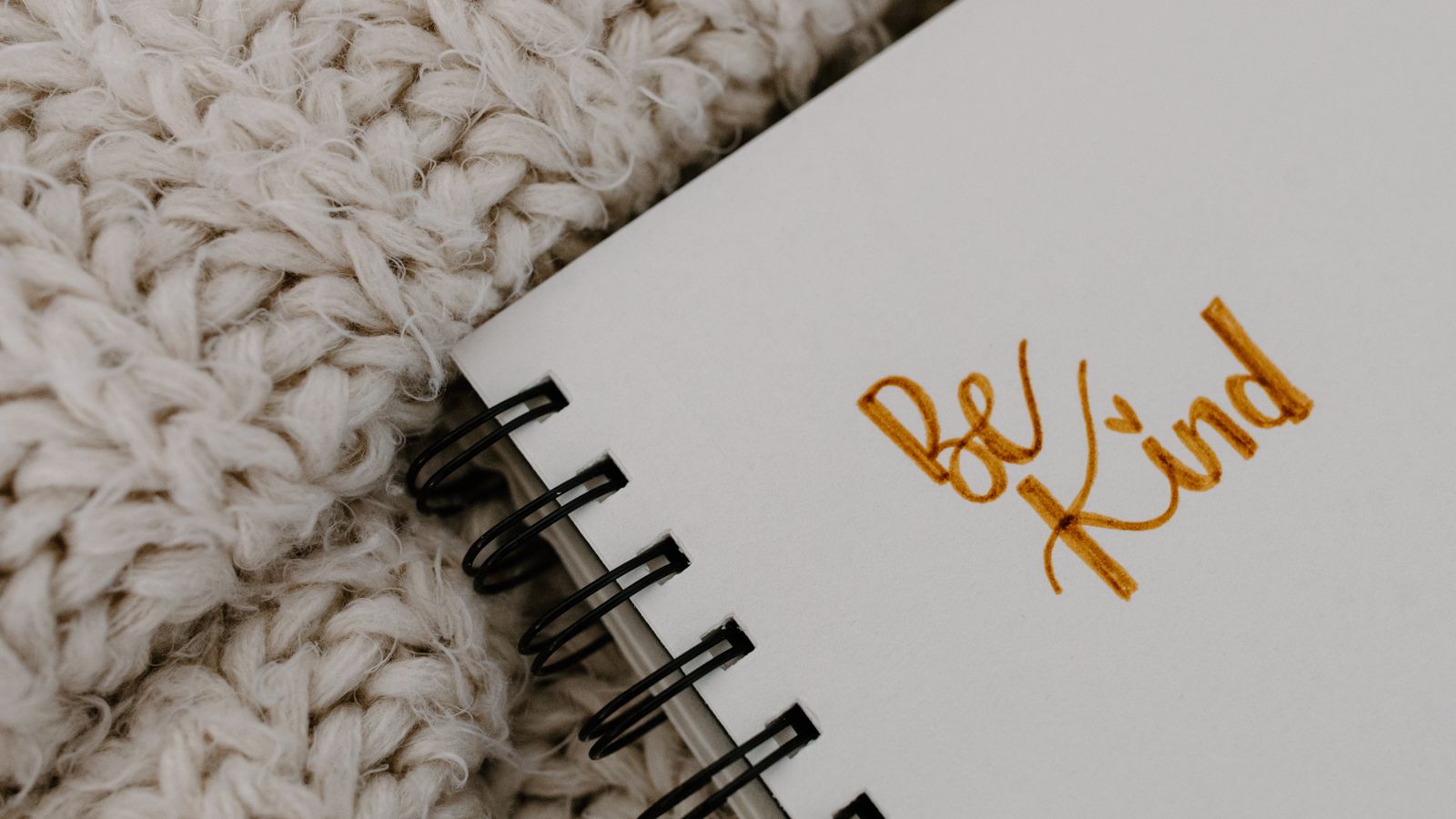 A handwritten “Be Kind” affirmation in a notebook resting on a warm knitted blanket