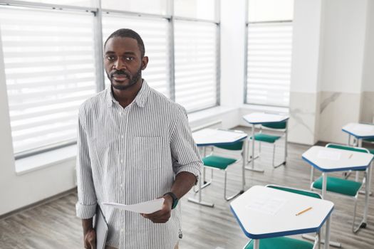 Adult male teacher standing in an empty classroom with papers, ready for a test.