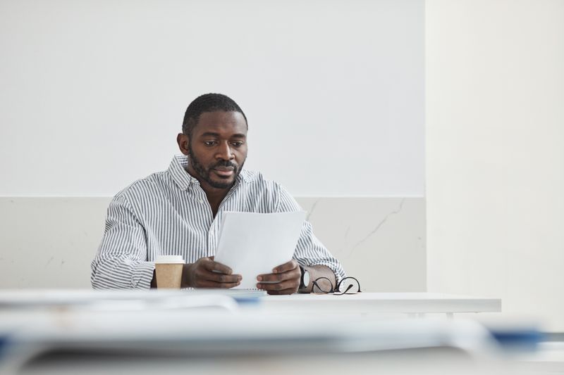 A Black man sits at a table with a coffee cup and reads a printed report closely. Reading your own lab results, starting with eGFR and uACR, is the first step in catching kidney disease early.