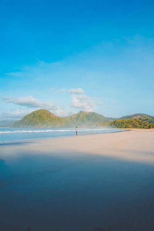Free Person walking on a tranquil beach with hills and blue sky, ideal for nature lovers. Stock Photo