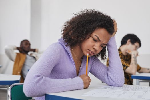 Young woman with curly hair in a classroom setting, thinking during an exam.