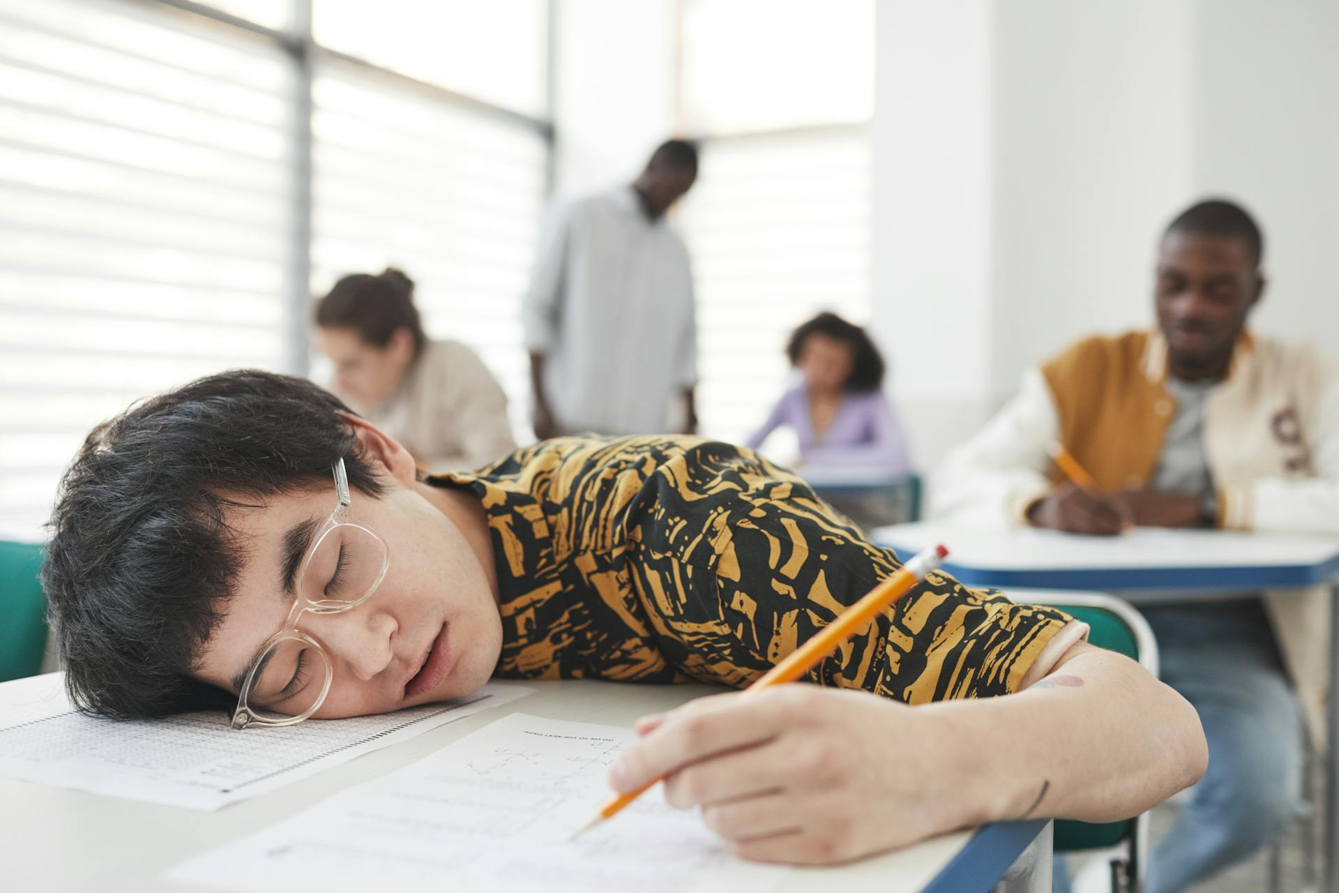 A student asleep on the exam paper in a classroom, appearing exhausted.