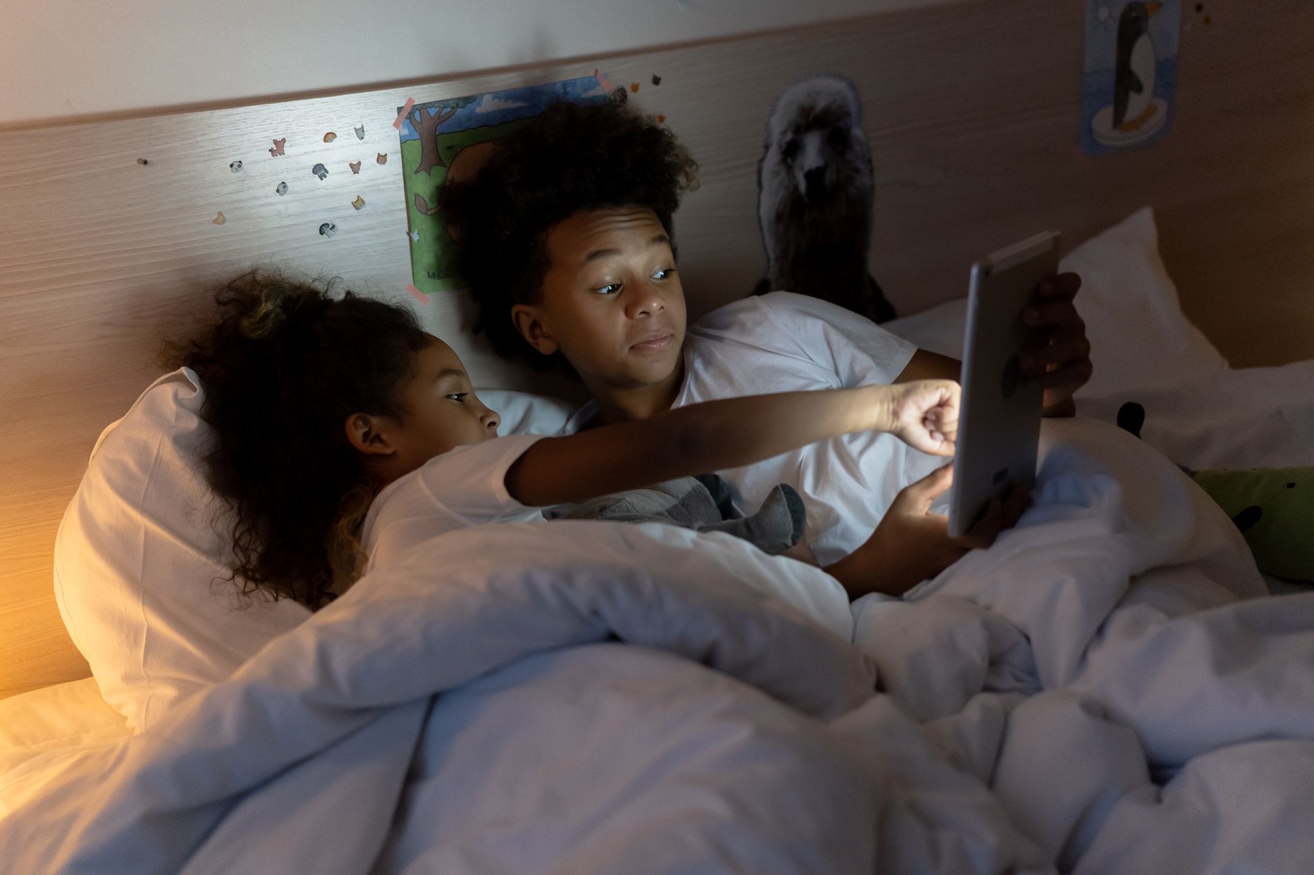 手機螢幕使用時間與數位健康:上限設定與例外的實務指南 Two siblings enjoying screen time together in bed during an evening indoors.