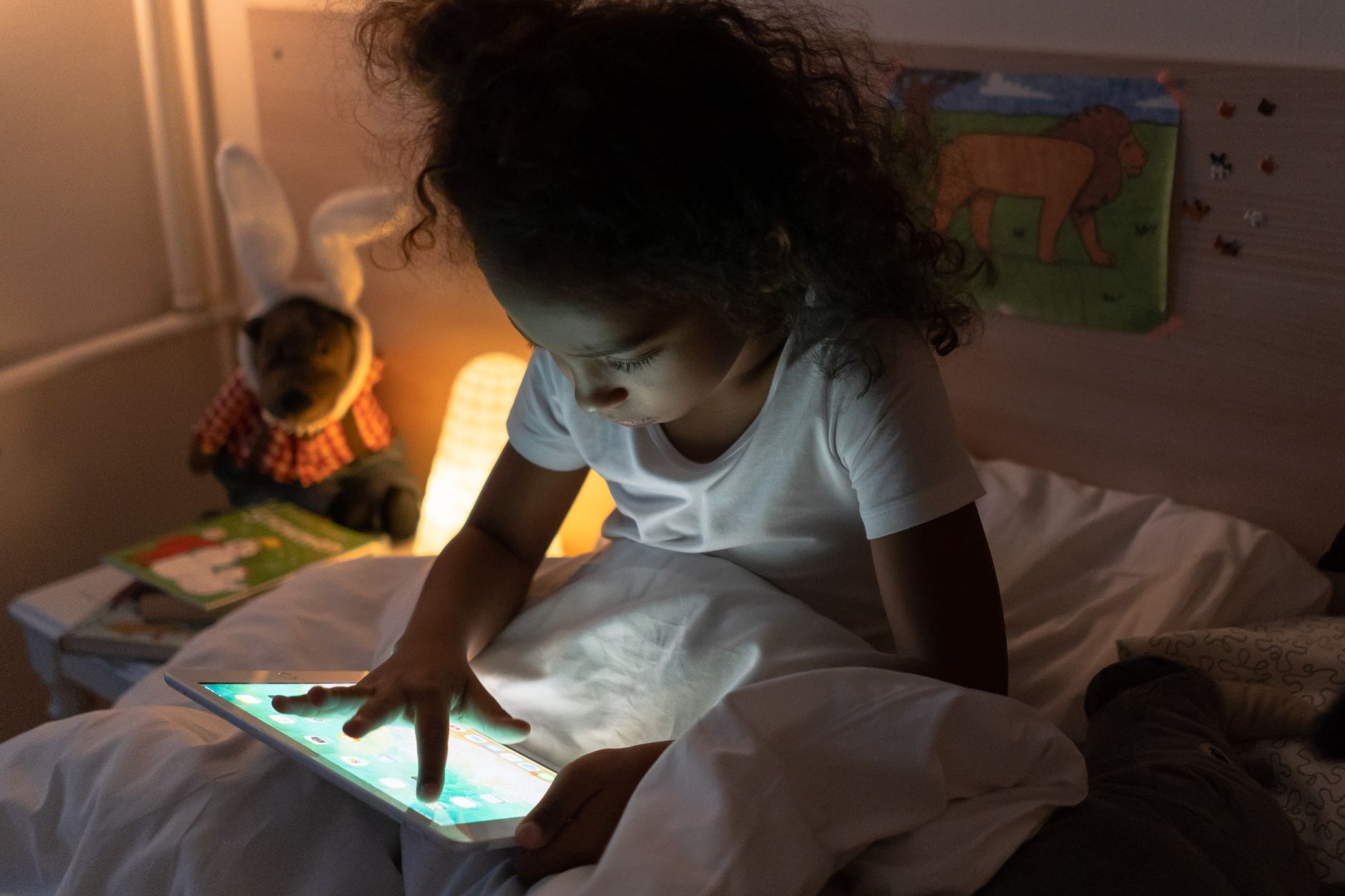 A young girl comfortably in bed, illuminated by a tablet's screen light at night.