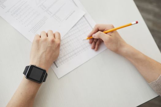 Unrecognizable student writing an exam using a pencil, watch visible on wrist.
