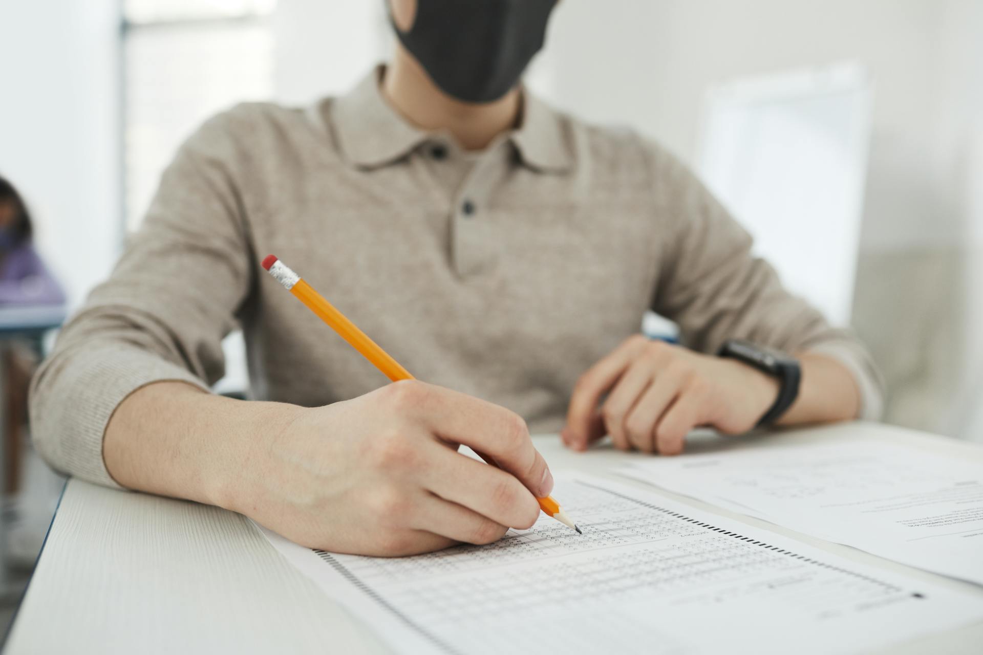 Person wearing a face mask writing a test at a desk, focusing on public health safety.