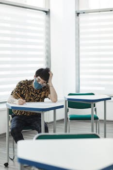 A student wearing a face mask sits alone at a desk in a classroom, focusing on an exam.