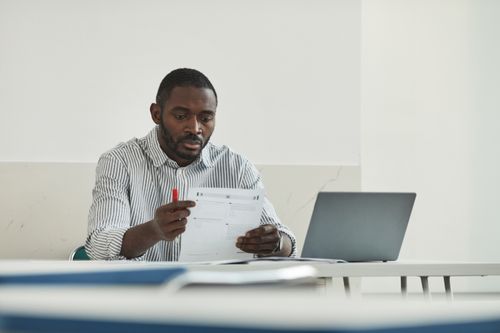 Free An adult man attentively grading test papers at his workspace with a laptop. Stock Photo