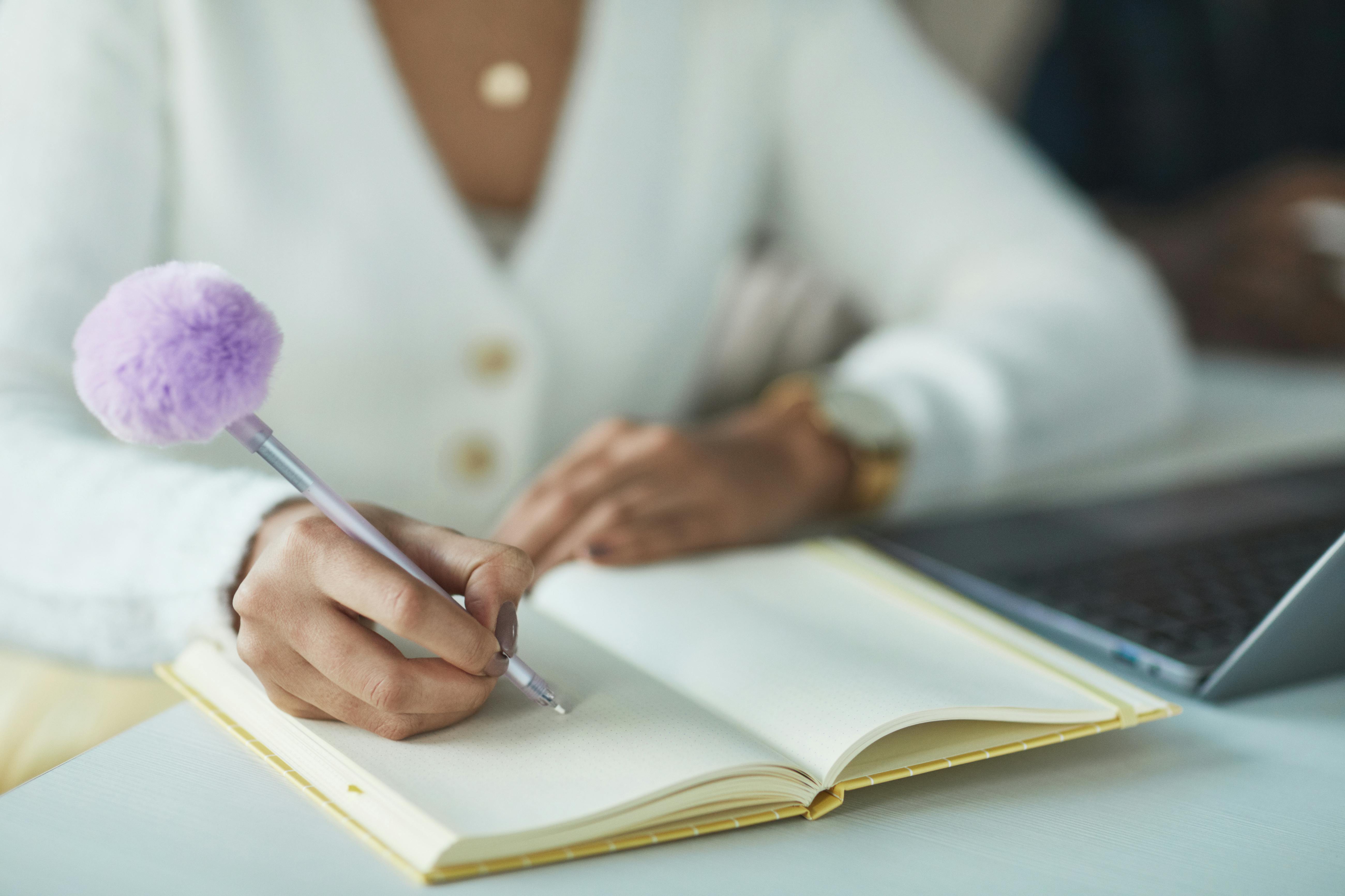 Close-up of a woman writing notes with a fluffy pen, indoors near a laptop.