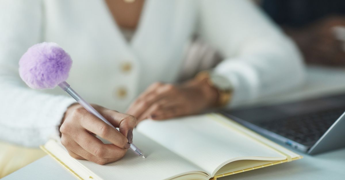 Close-up of a hand writing in a journal beside a laptop
